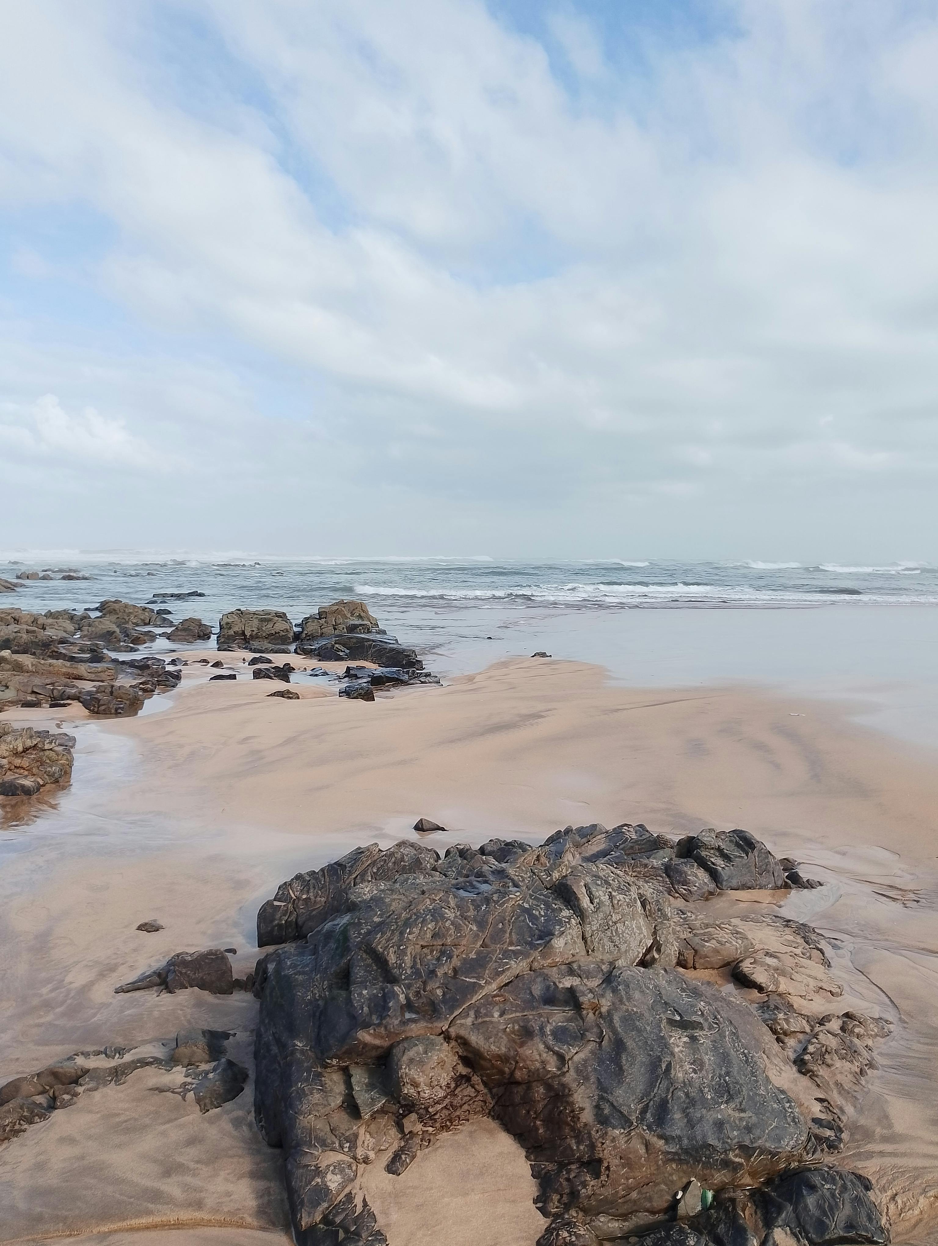 A serene beach scene with shallow waves lapping at scattered dark rocks on wet sand, under a partly cloudy sky, with the ocean extending to the horizon.