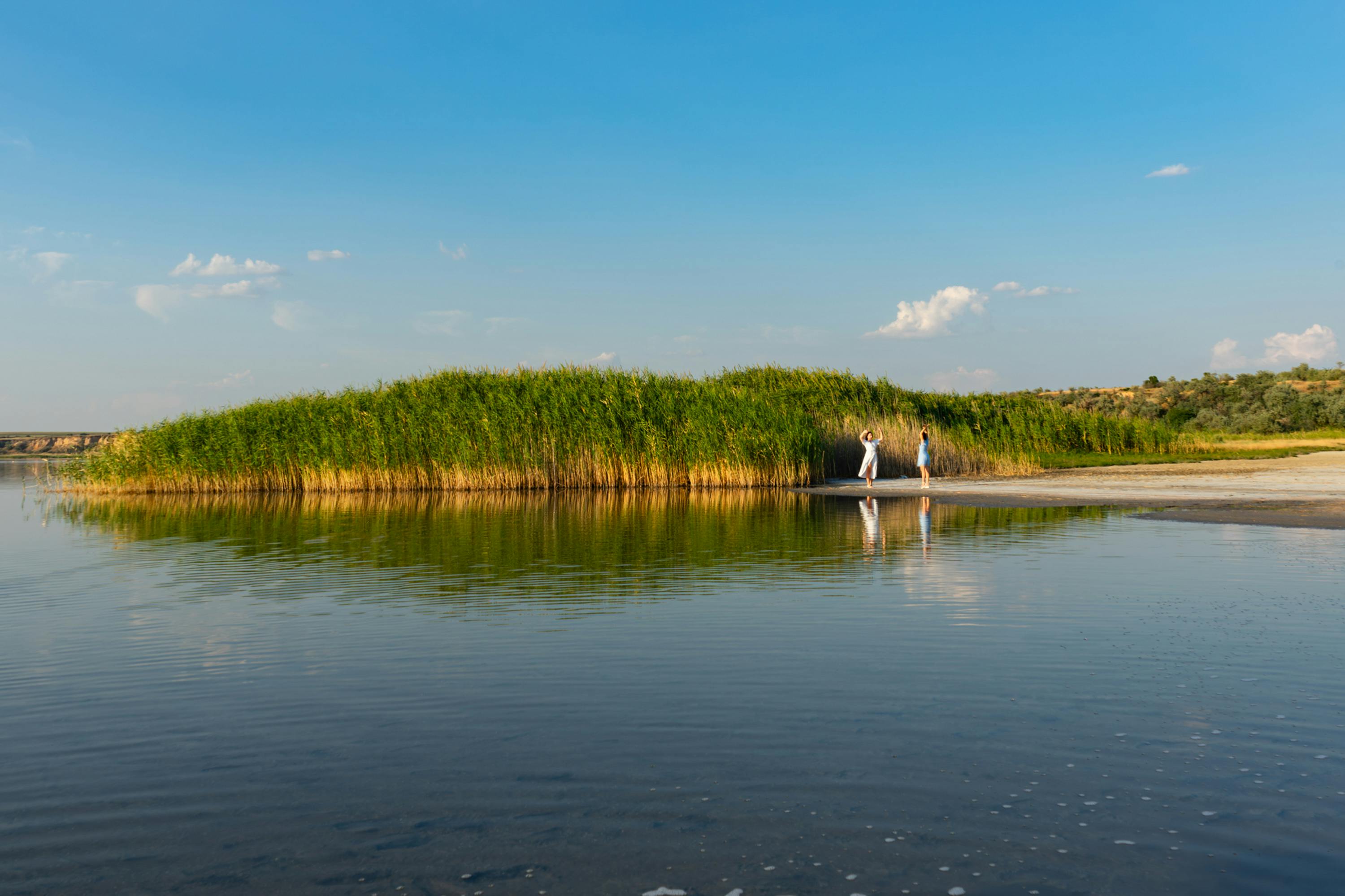 Two people standing near a lush green island by a serene lake under a clear blue sky.