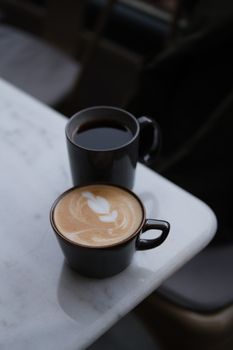 A close-up of a latte with intricate art next to a black coffee on a marble table setting.