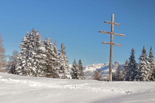 Scenic winter landscape with a wooden cross, snowy trees, and mountains in Trentino-Alto Adige.
