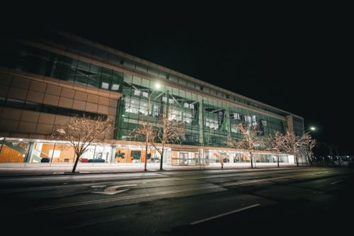 Free Contemporary glass building facade illuminated with lights at night, featuring leafless trees and empty street in foreground. Stock Photo