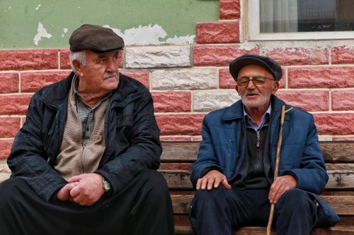 Free Two senior men in casual clothing sitting on a wooden bench against a brick wall. Stock Photo