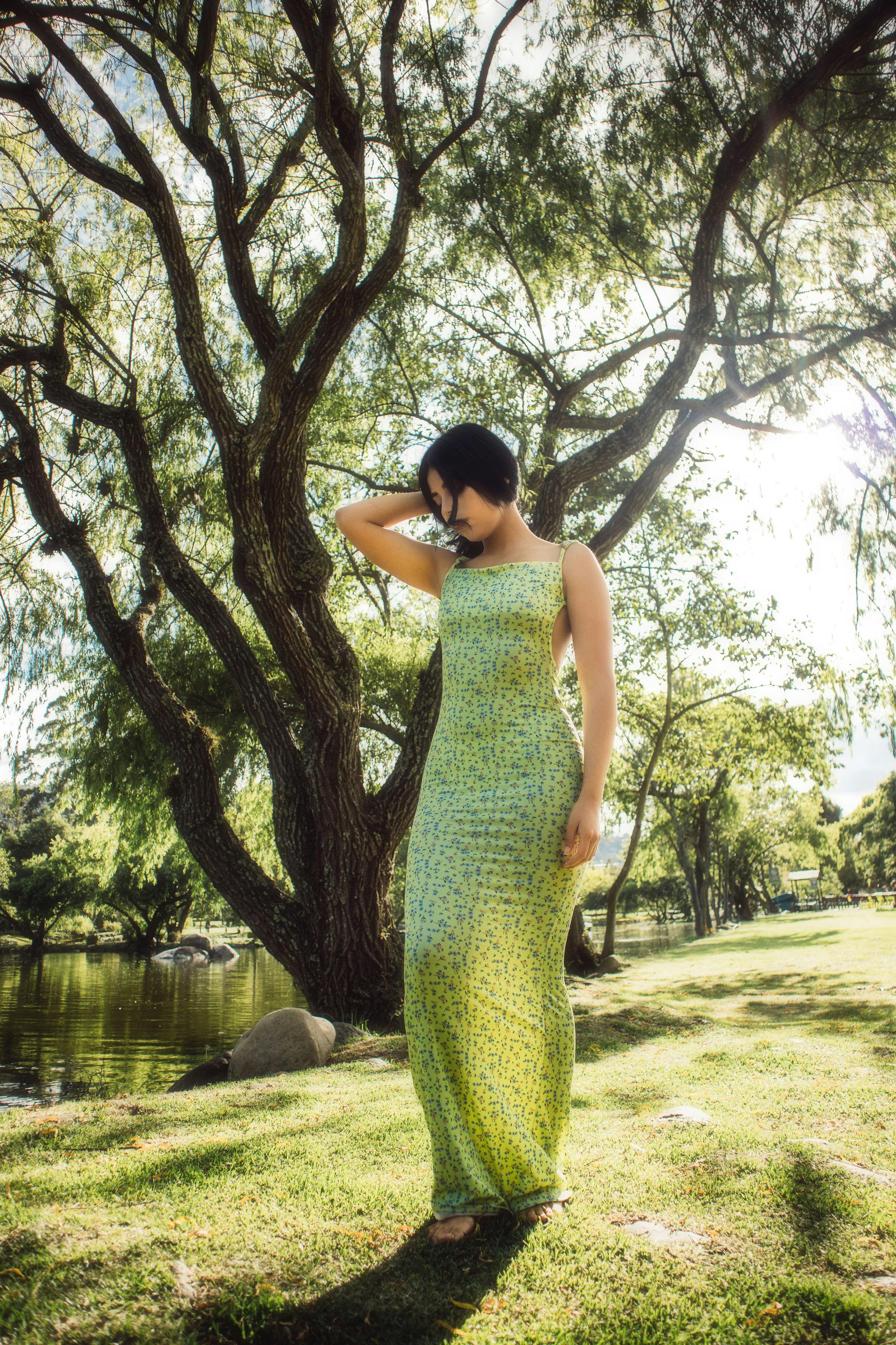 Elegant woman in green dress stands in a sunlit park under a large tree, evoking a serene summer vibe.