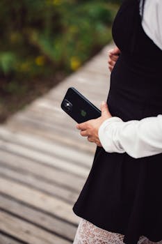 Pregnant woman in black dress holds smartphone, walking on wooden path outdoors.