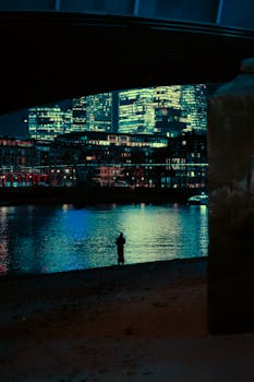 A solitary figure stands by the riverbank under a bridge with neon reflections of London's cityscape.
