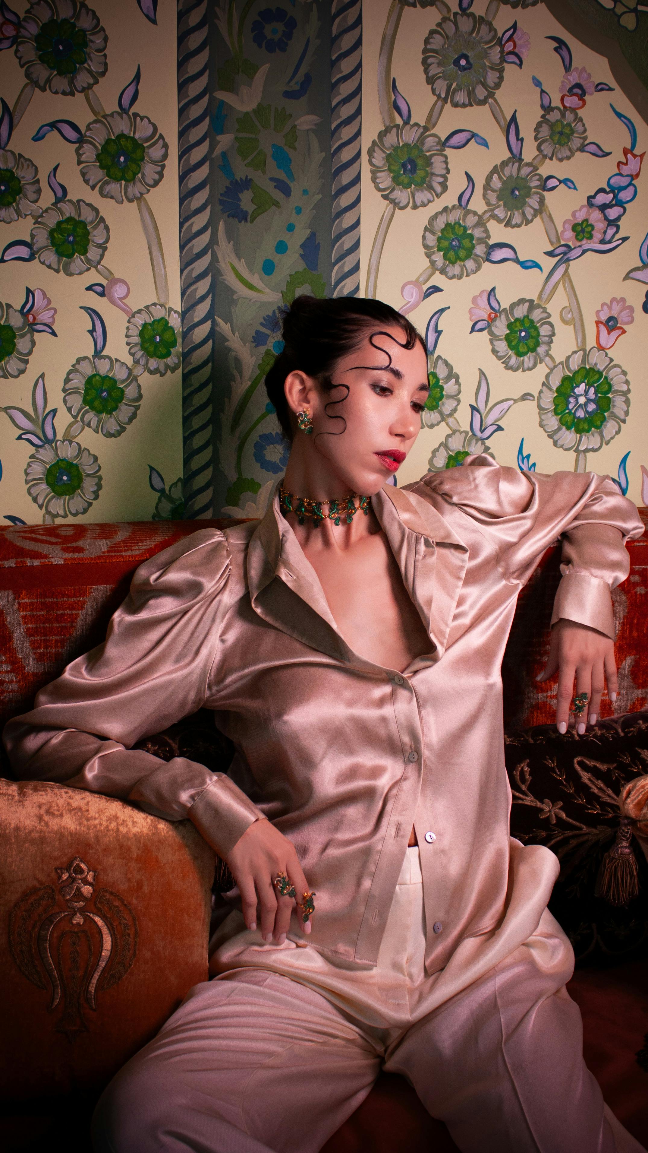 Stylish woman in silk outfit poses on ornate furniture with floral wallpaper backdrop.
