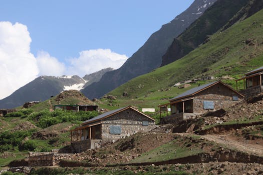 Stone cabins in a lush valley setting against a mountainous backdrop under a blue sky.