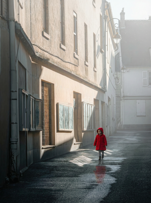 Free A child in a red coat walks down a misty street, casting reflections on wet pavement. Stock Photo