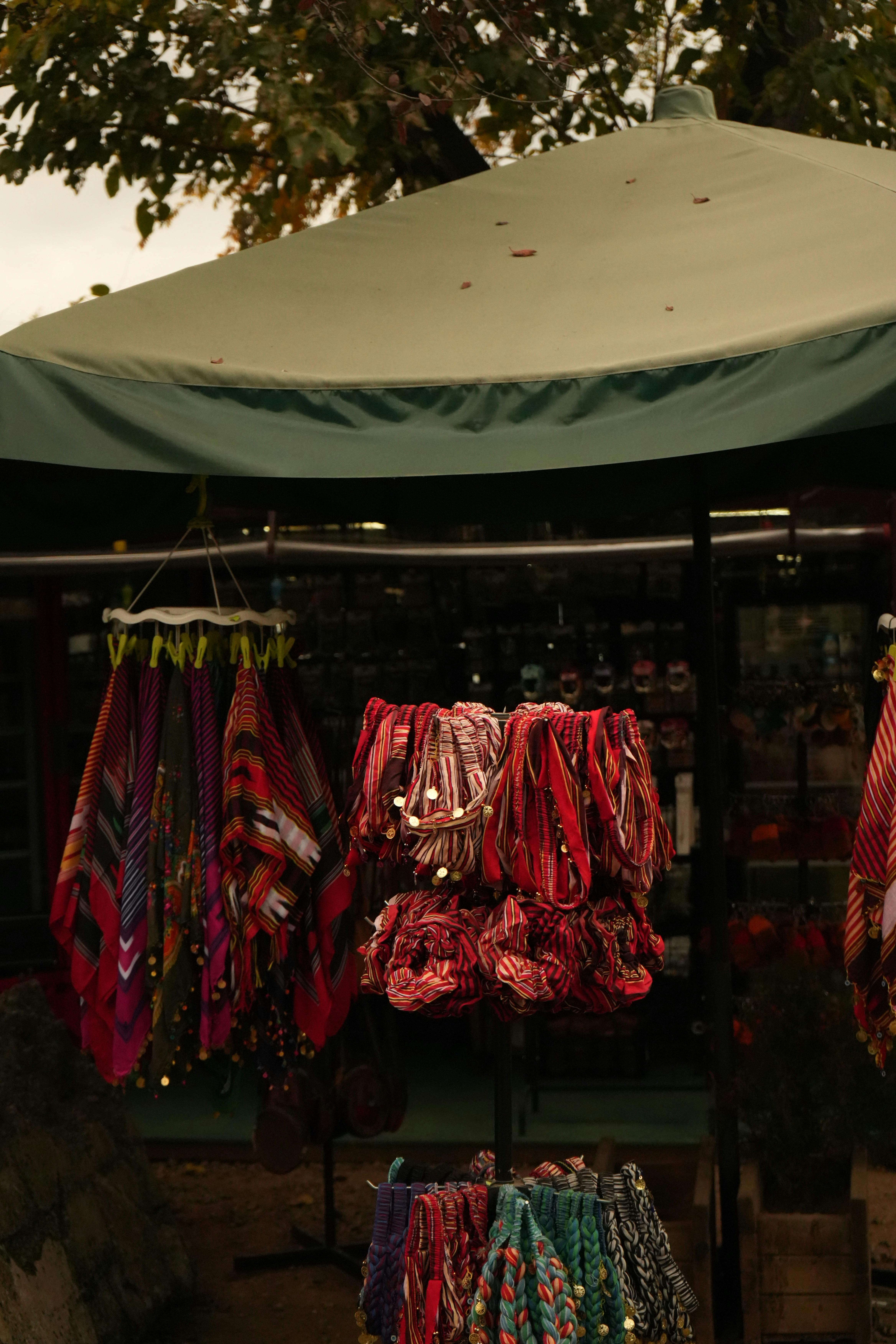Vibrant patterned scarves hang at a bustling outdoor market stall, under a green canopy.