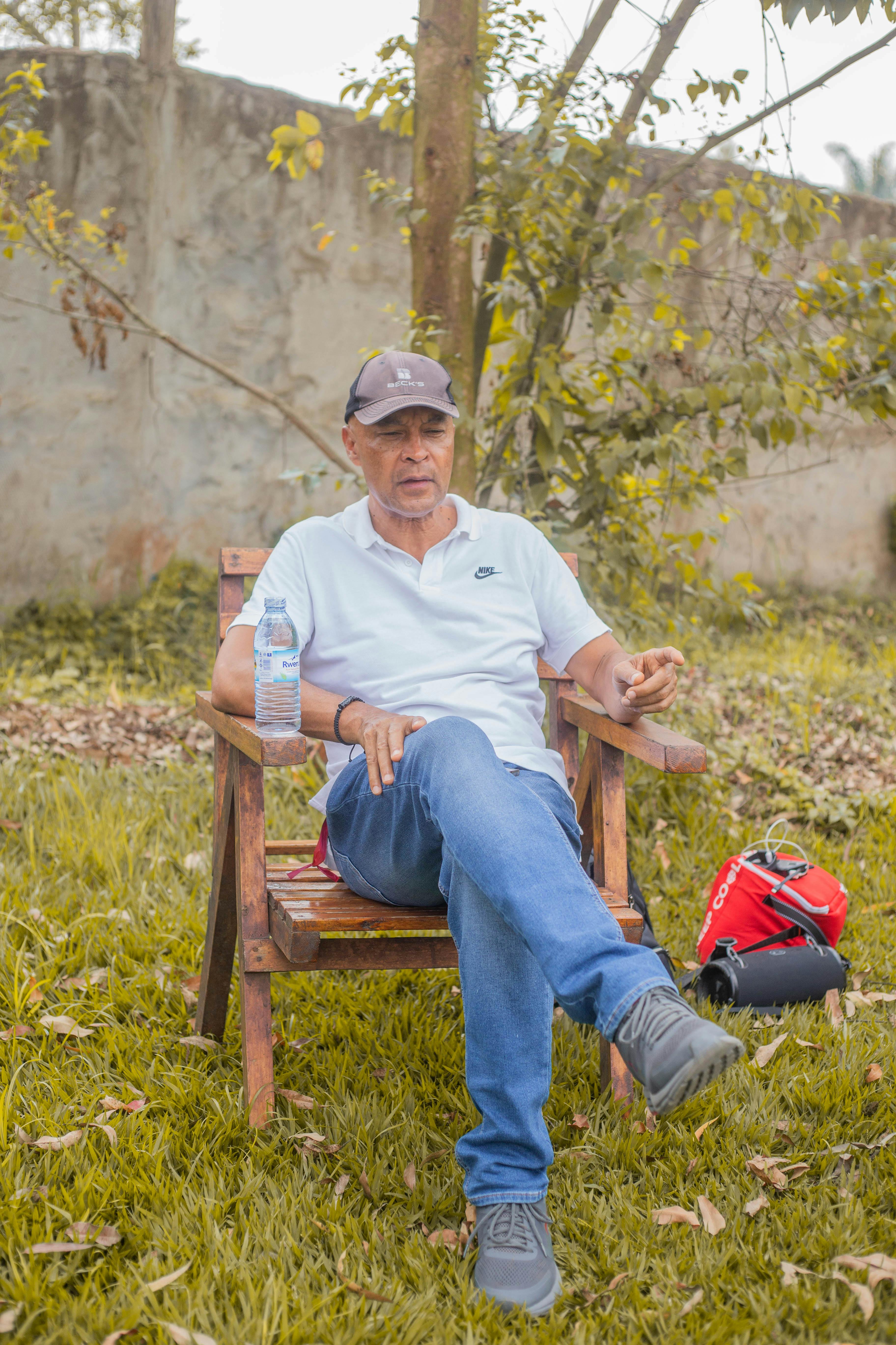Senior man sitting on wooden chair outdoors with a camera and red bag, enjoying a peaceful moment.