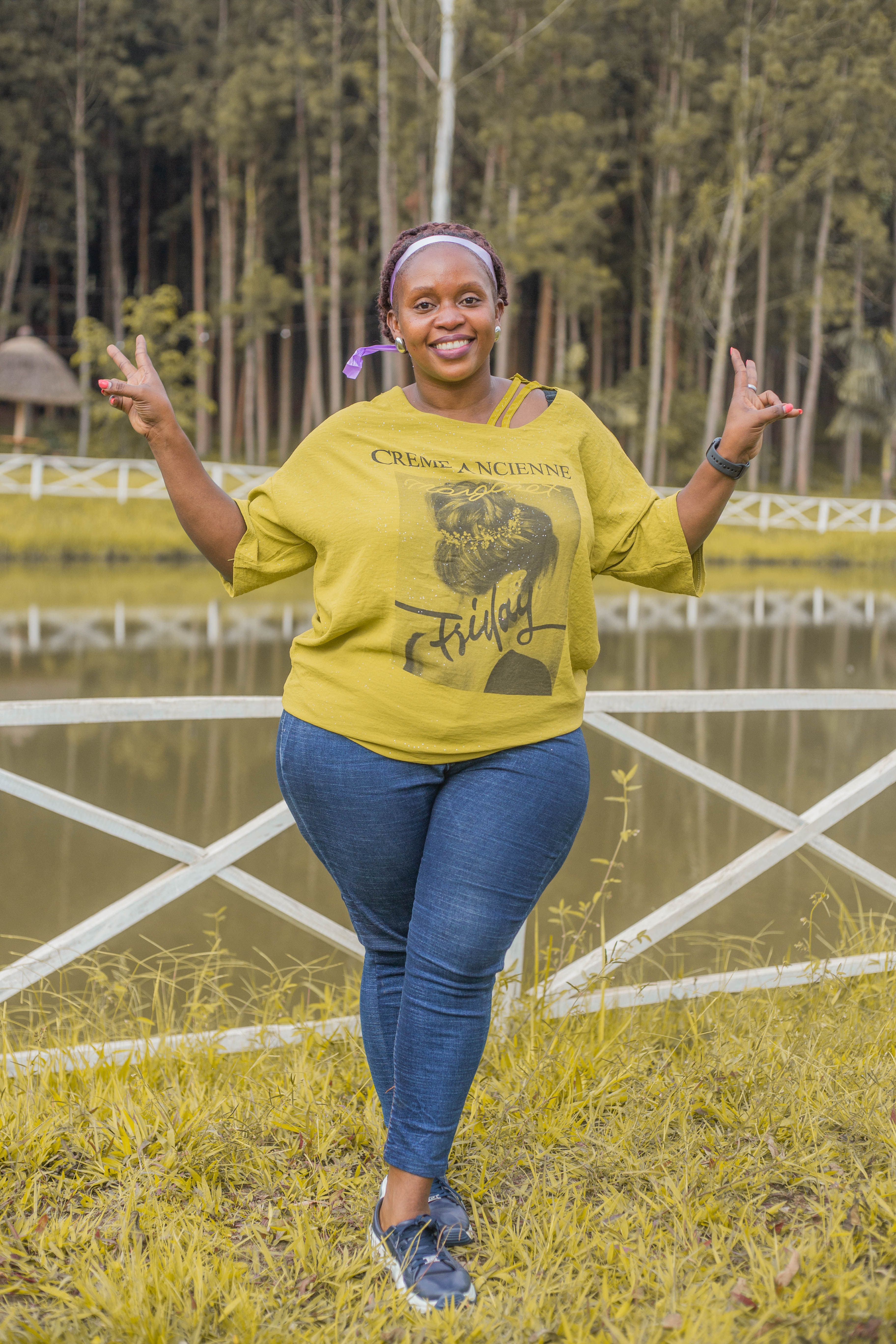 Smiling woman posing with peace signs near a serene lake, surrounded by lush forest.