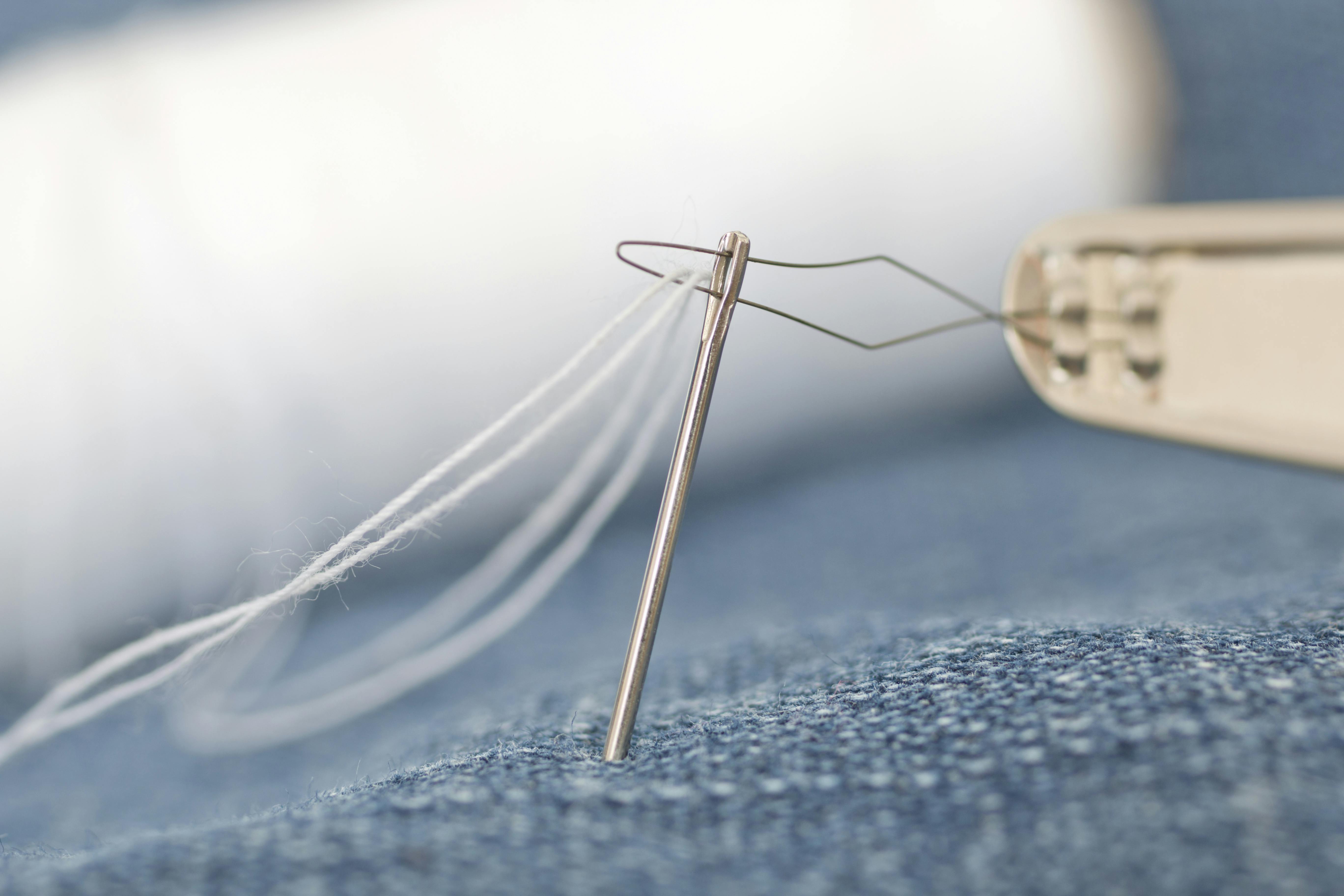 Detailed macro shot of a sewing needle threading white thread through denim fabric.