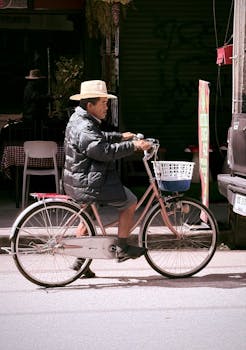 Adult male riding a bicycle on an urban street during the day, wearing a jacket and hat.