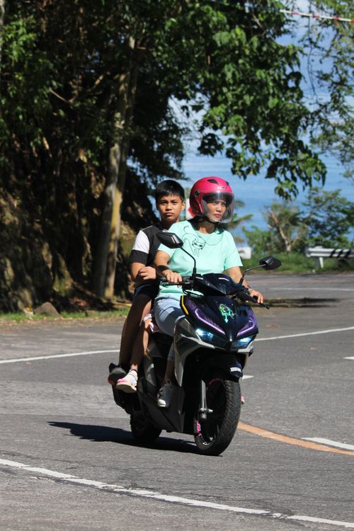 Free Two individuals riding a scooter on a sunny day along a scenic road. Stock Photo