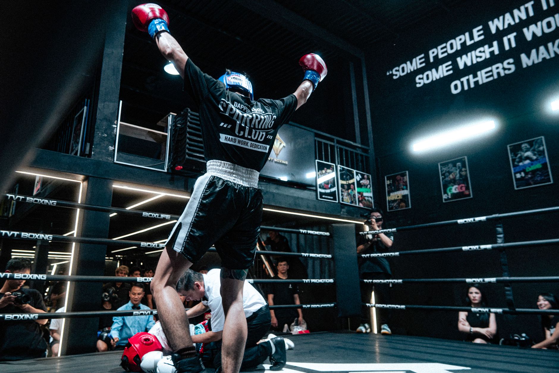 A victorious boxer raises his arms in triumph in a dimly lit boxing ring after a match.
