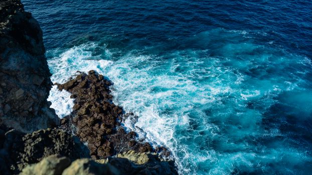Aerial view of ocean waves crashing against rugged cliffs with vibrant blue water.