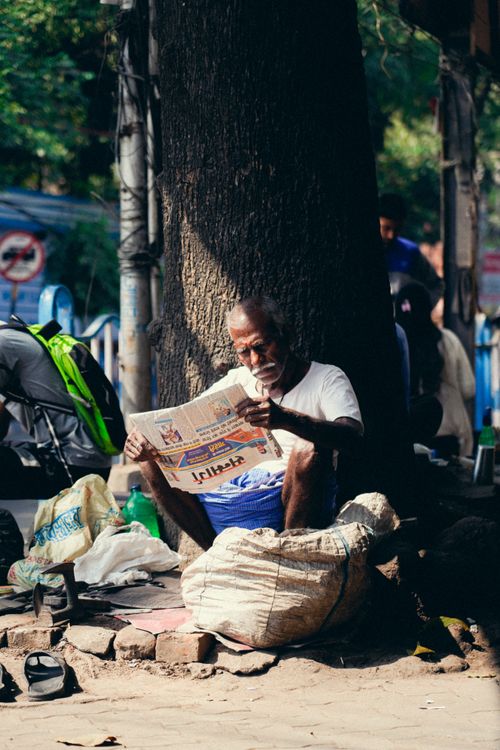 Free A senior man reads a newspaper under the shade of a tree on a bustling city street, surrounded by bags. Stock Photo