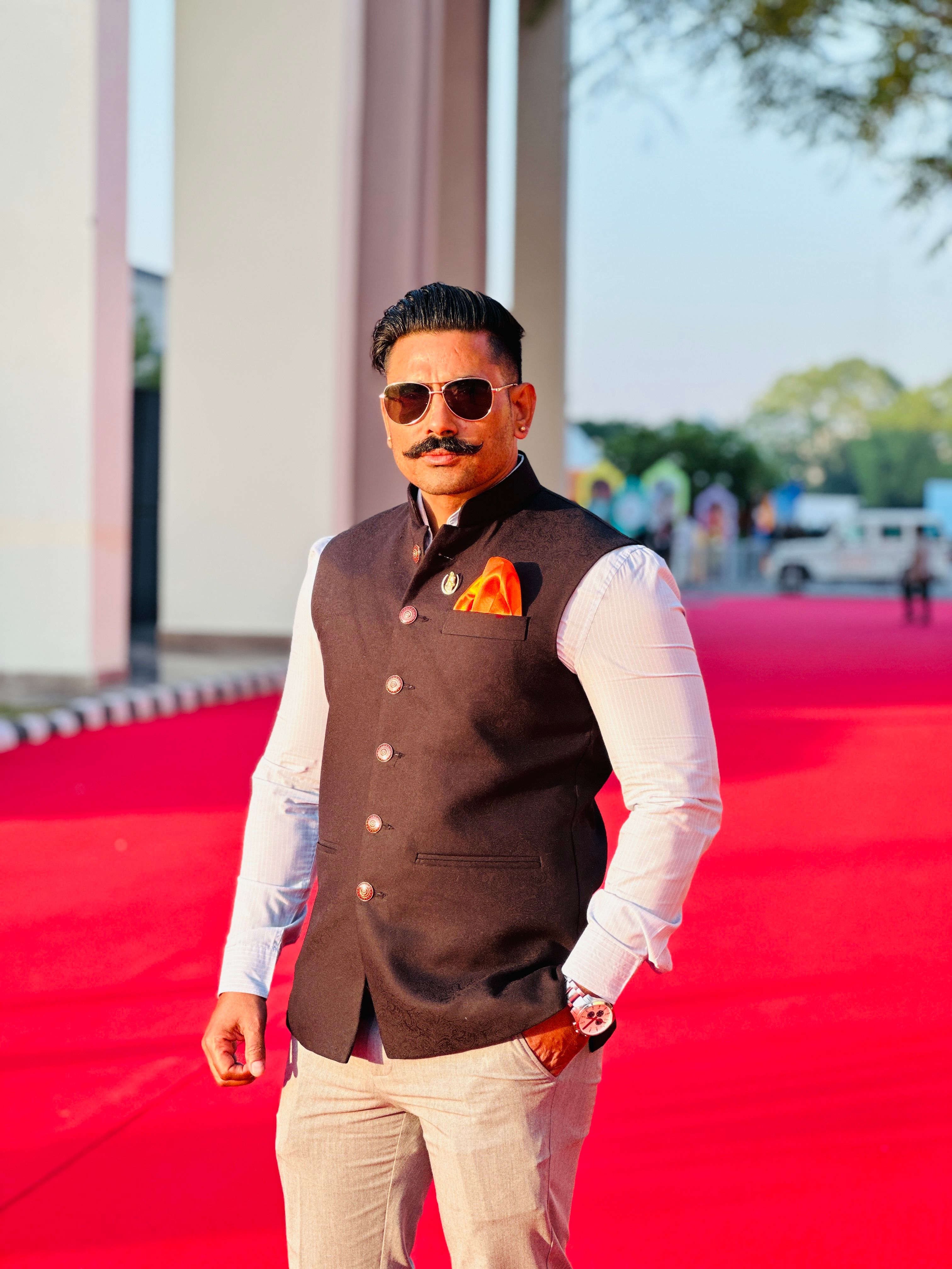 Confident man in formal attire standing on a red carpet outdoors with a sunny atmosphere.