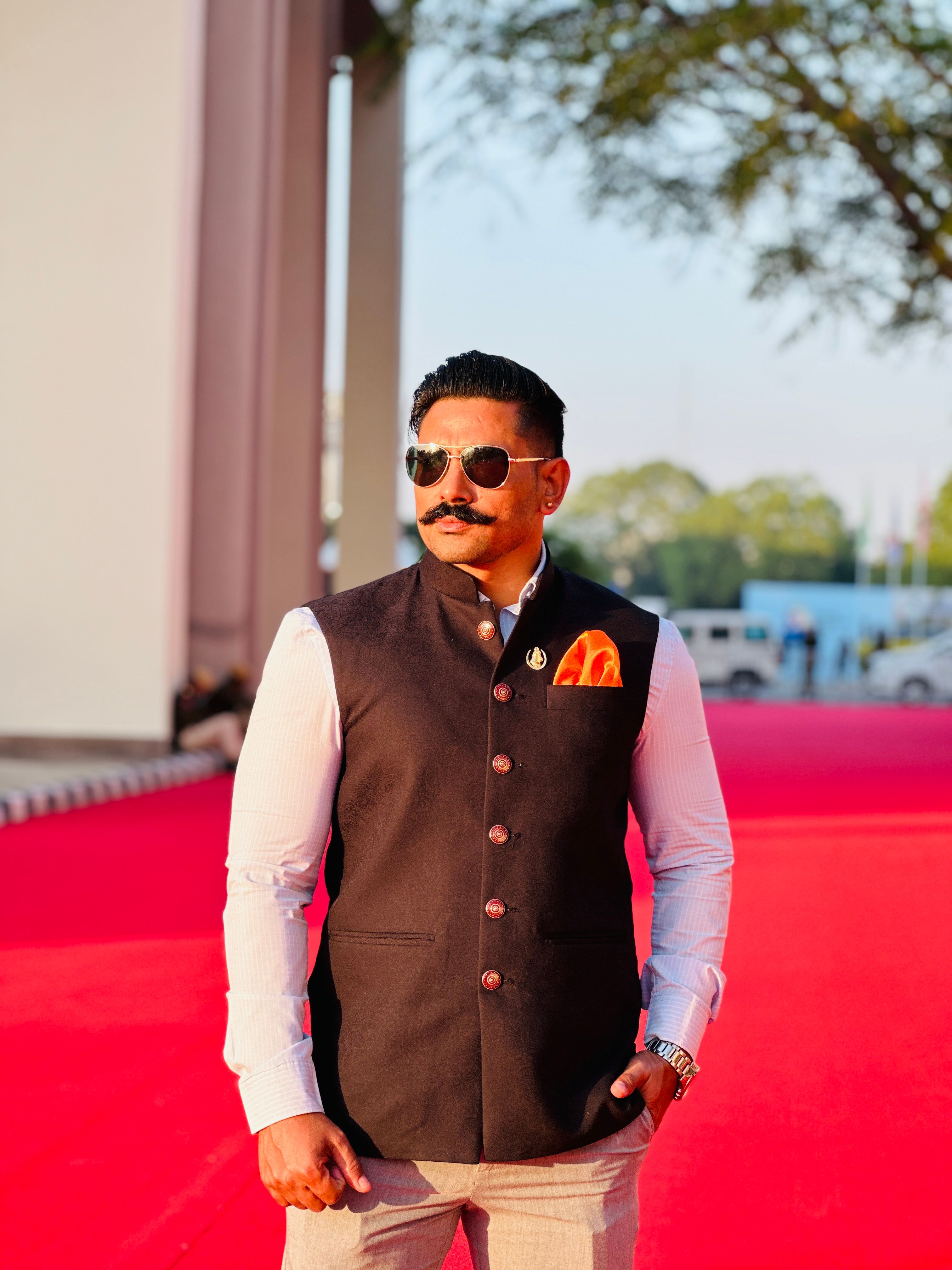 Man in stylish attire with sunglasses standing on a red carpet outdoors.