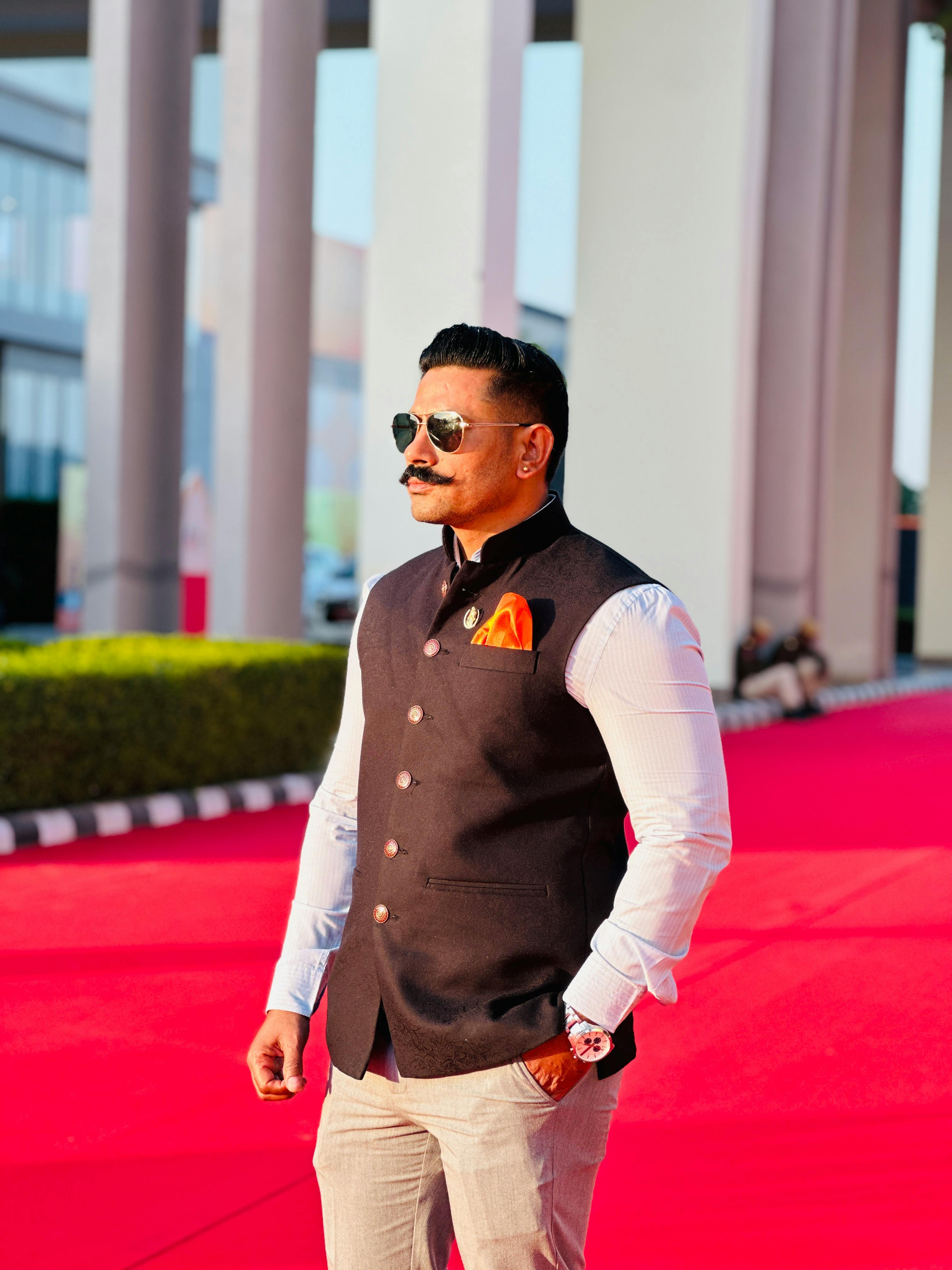 Stylish man wearing sunglasses and traditional attire on a red carpet outdoors.