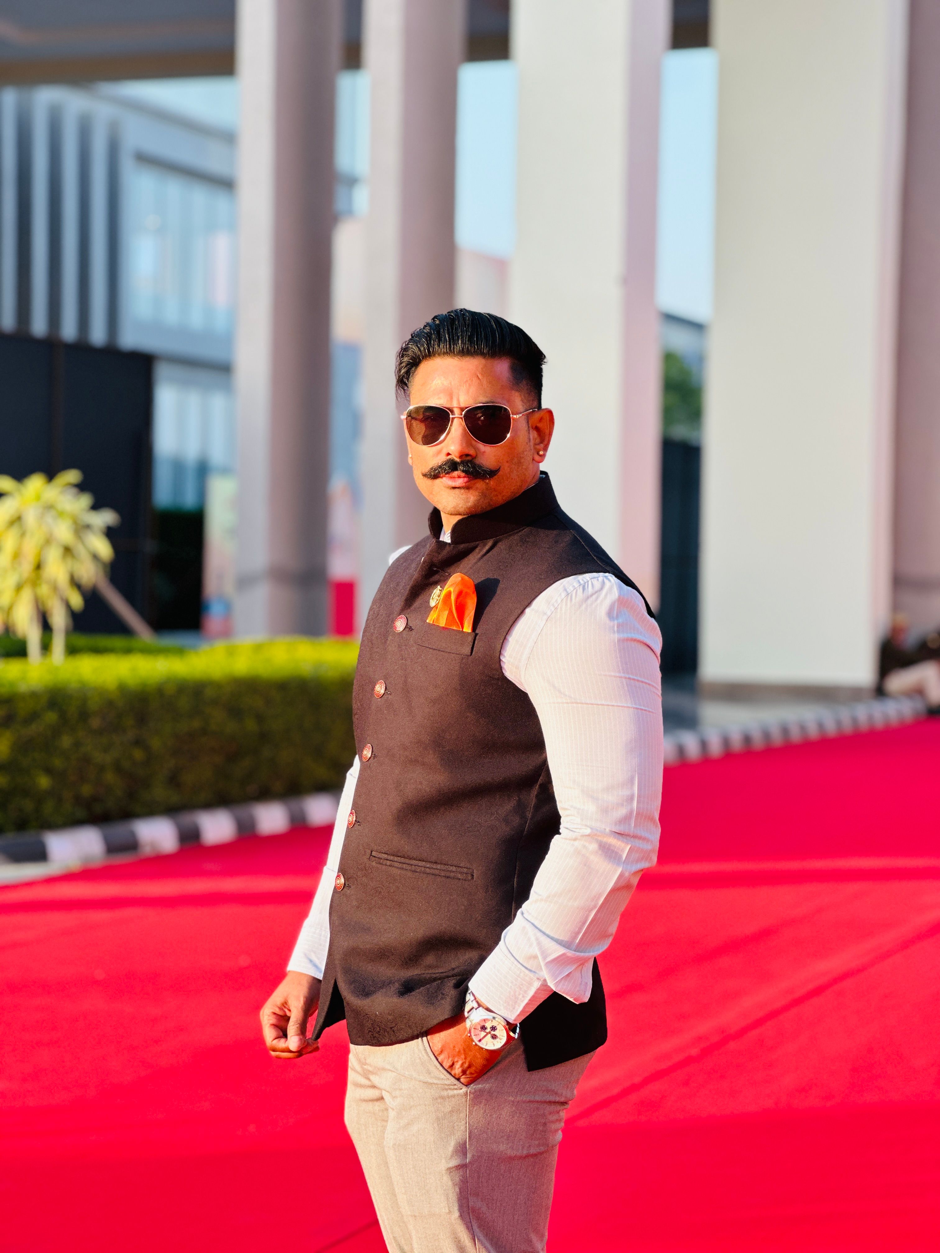 Confident man with sunglasses standing on red carpet in outdoor setting.