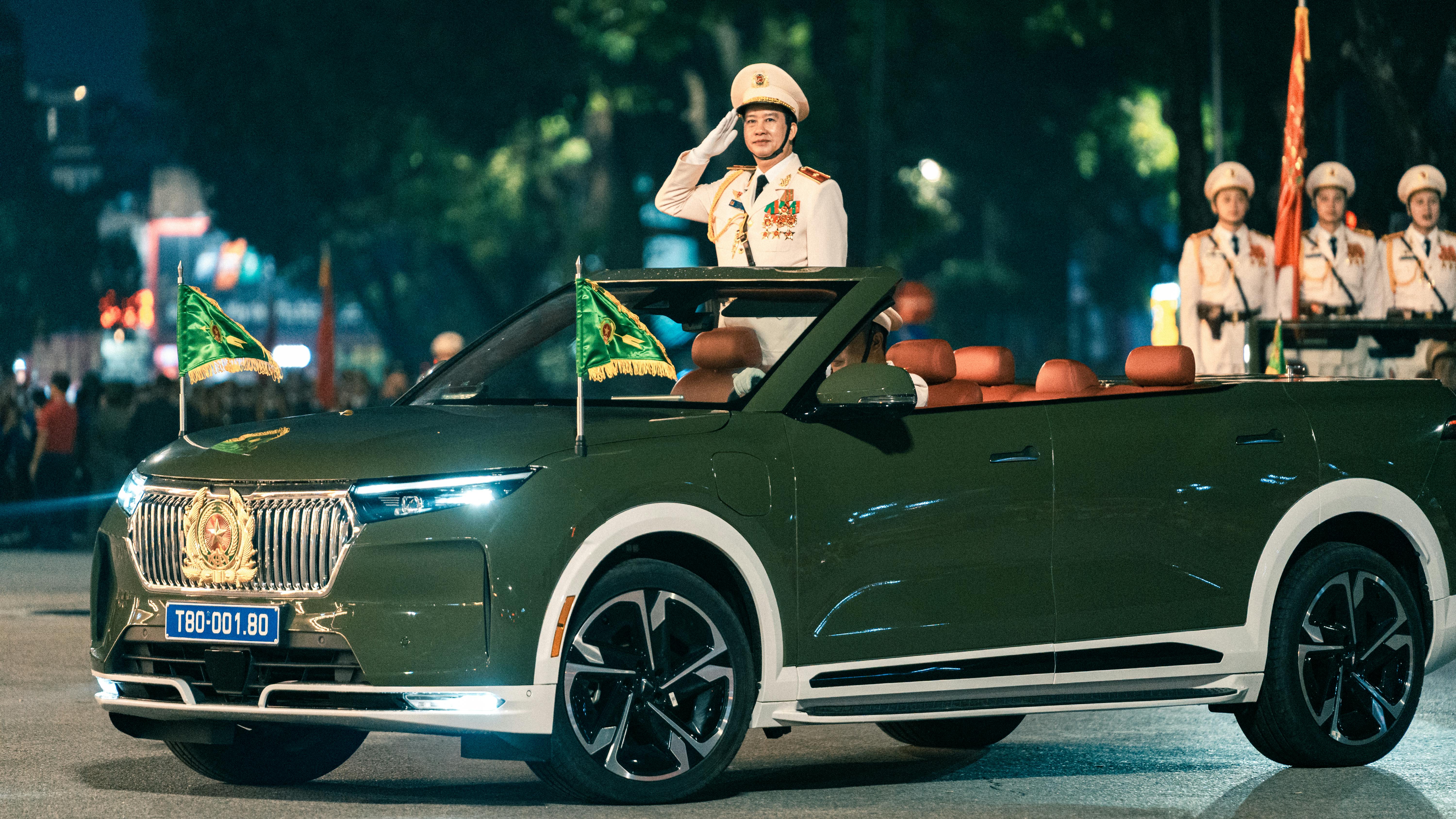 A military officer saluting during a parade in Hanoi with a convertible car and flags.