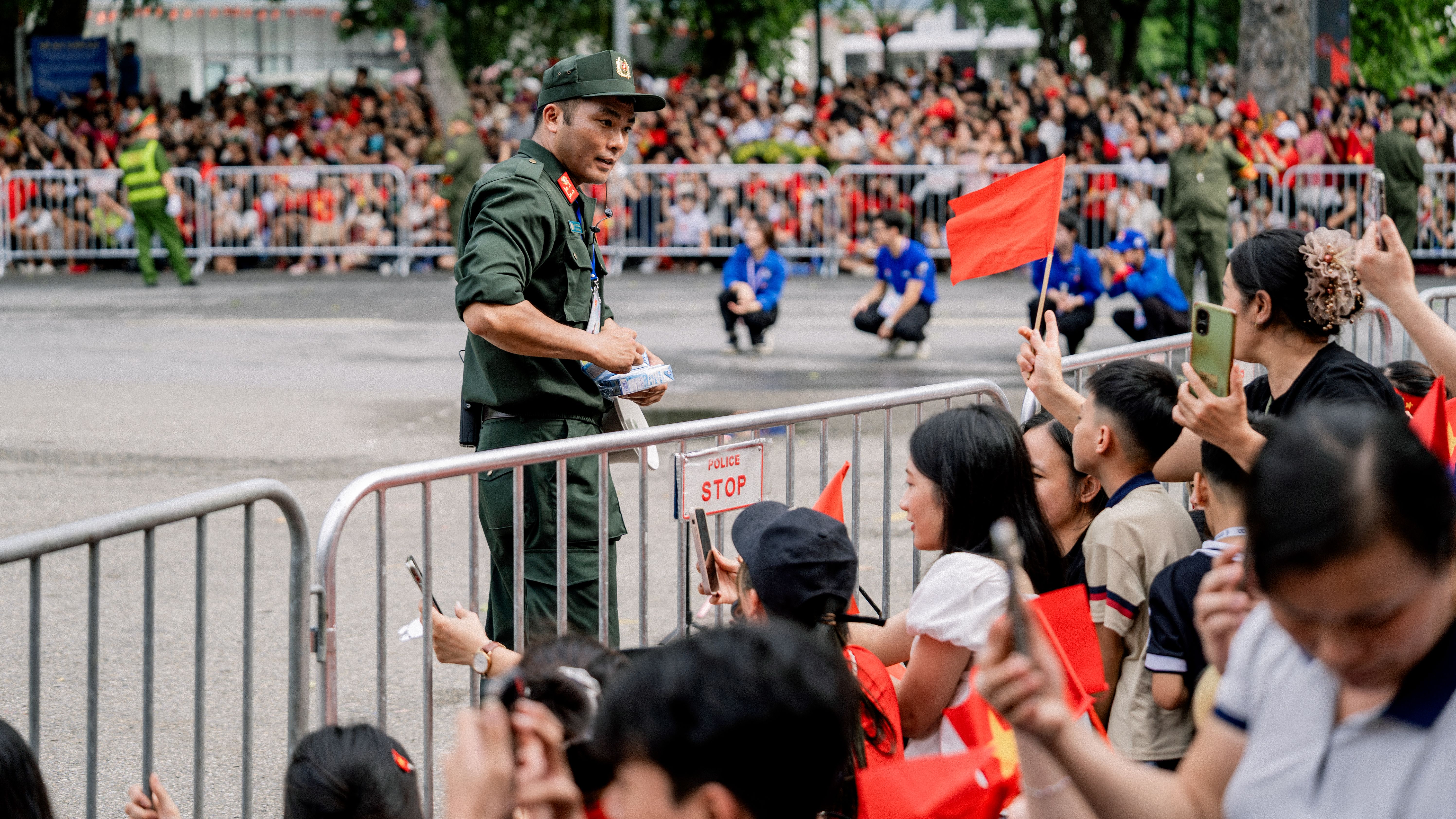 A crowd gathers at a parade in Hà Nội with people holding flags and police providing security.