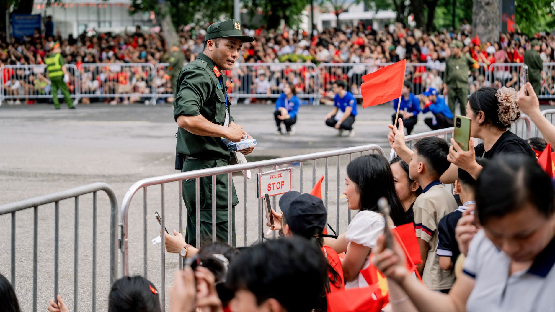 A crowd gathers at a parade in Hà Nội with people holding flags and police providing security.