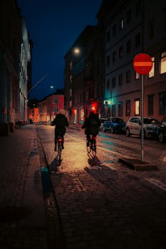 Two cyclists pedal through a dimly lit urban street at night, evoking a moody and cinematic atmosphere.