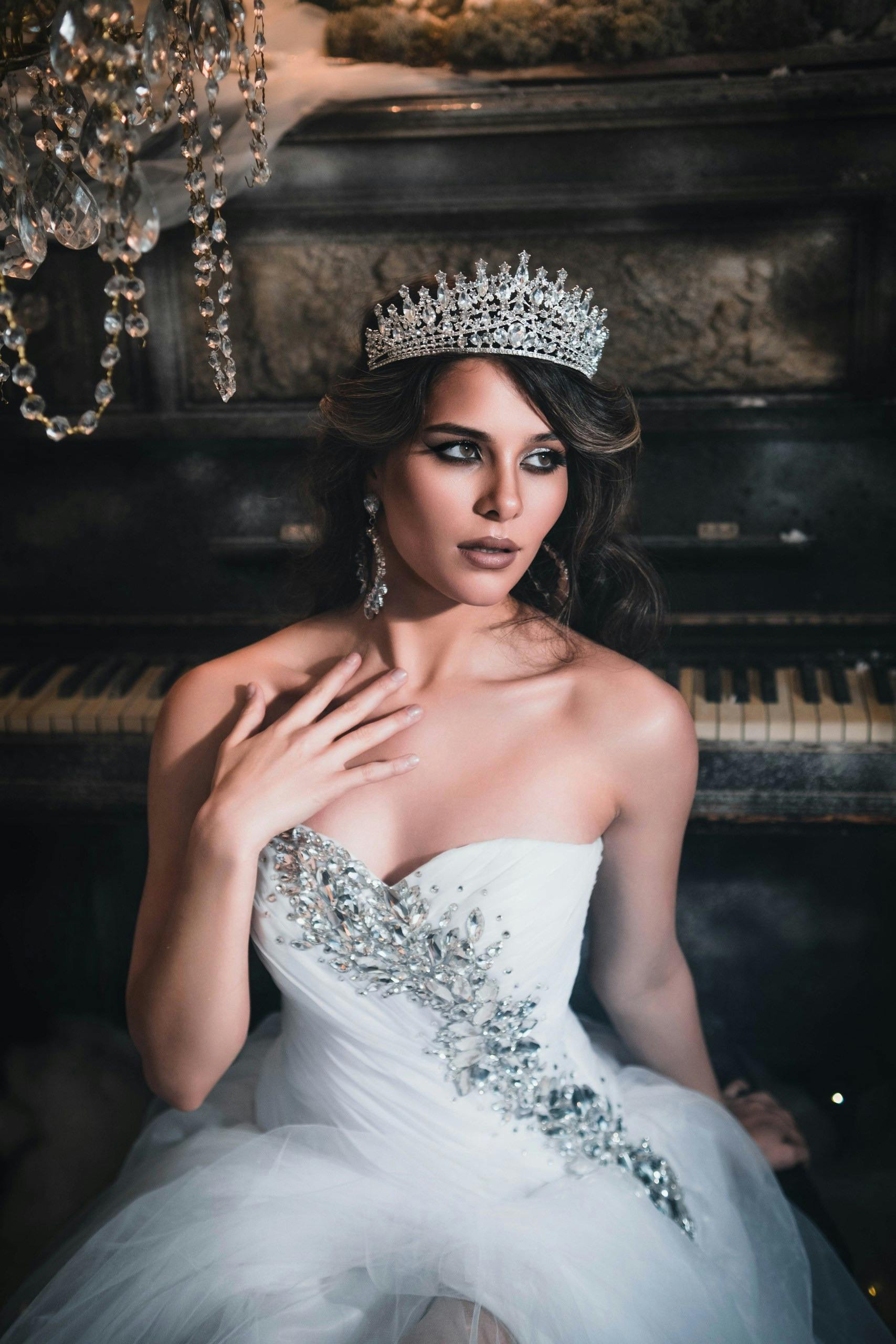 Stunning bride in an embellished white dress with a tiara, seated by a piano.