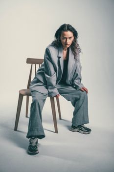 Stylish portrait of a woman in an oversized suit sitting confidently on a wooden chair.