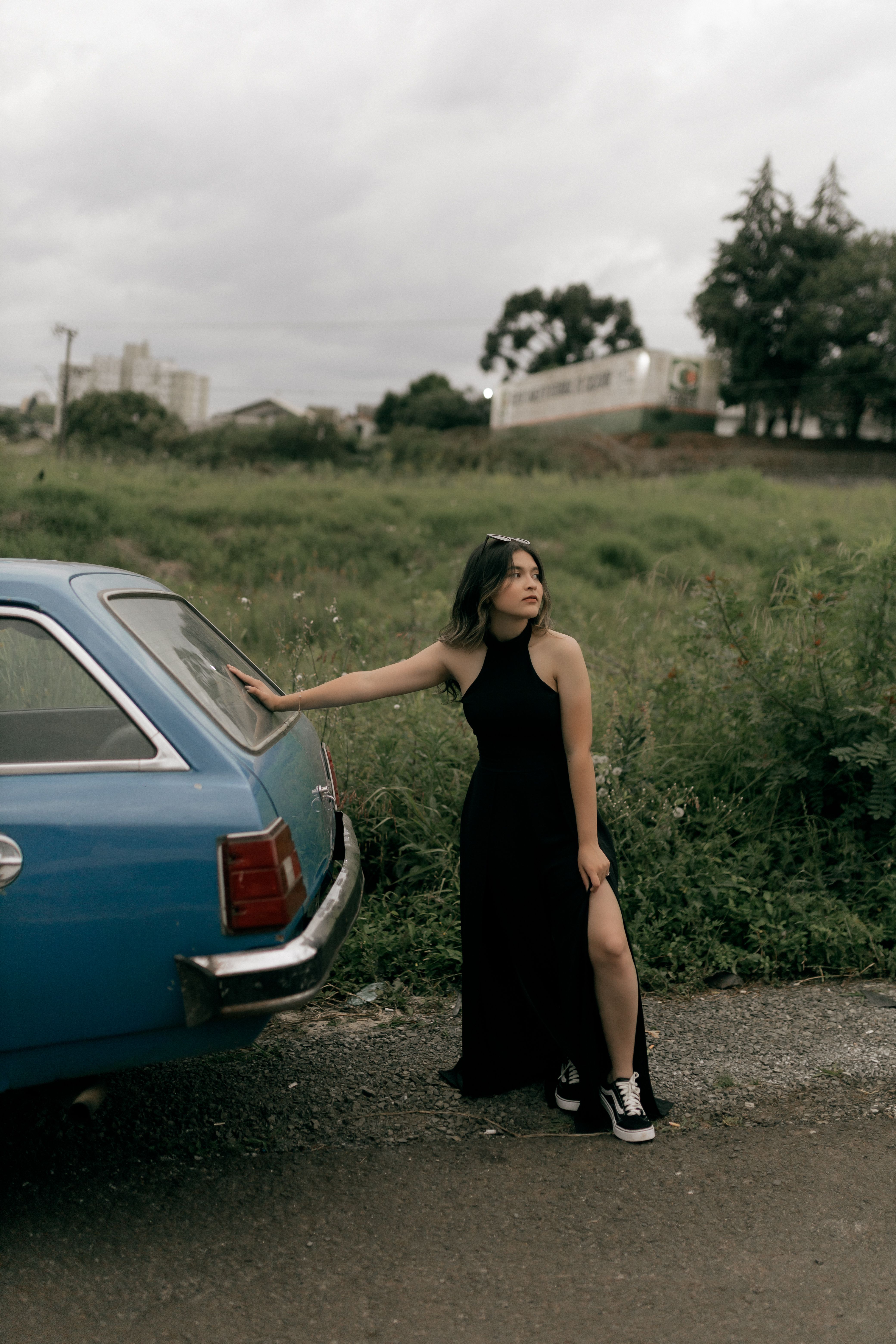 A young woman in a black dress stands by a vintage blue car, outdoors on a cloudy day.