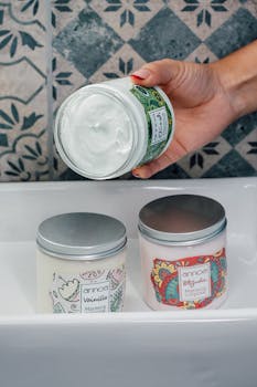 Hand holding a jar of skincare cream over two other cream jars on a patterned bathroom tile backdrop.