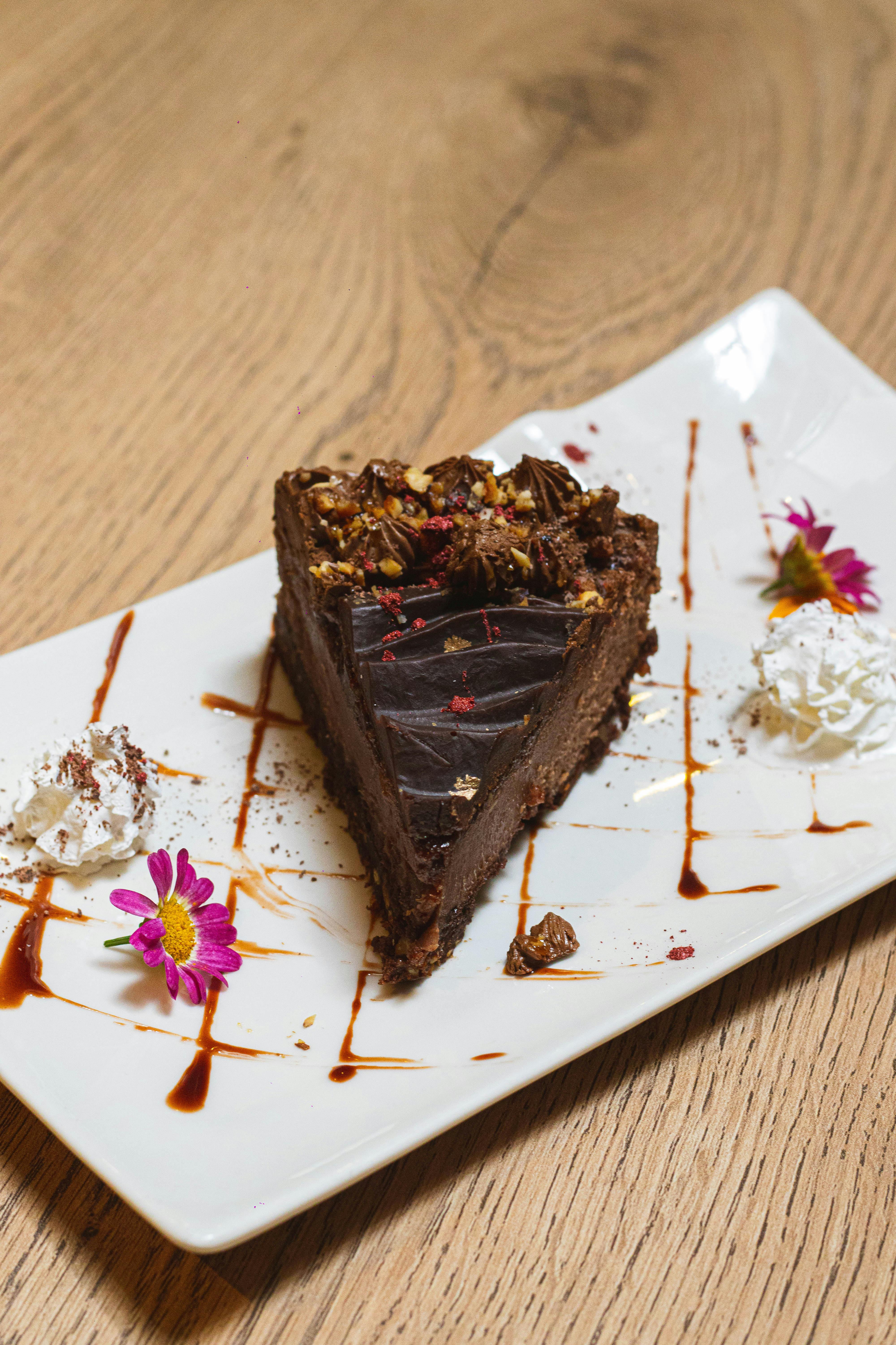 A rich chocolate cake slice garnished with whipped cream and edible flowers on a wooden table.