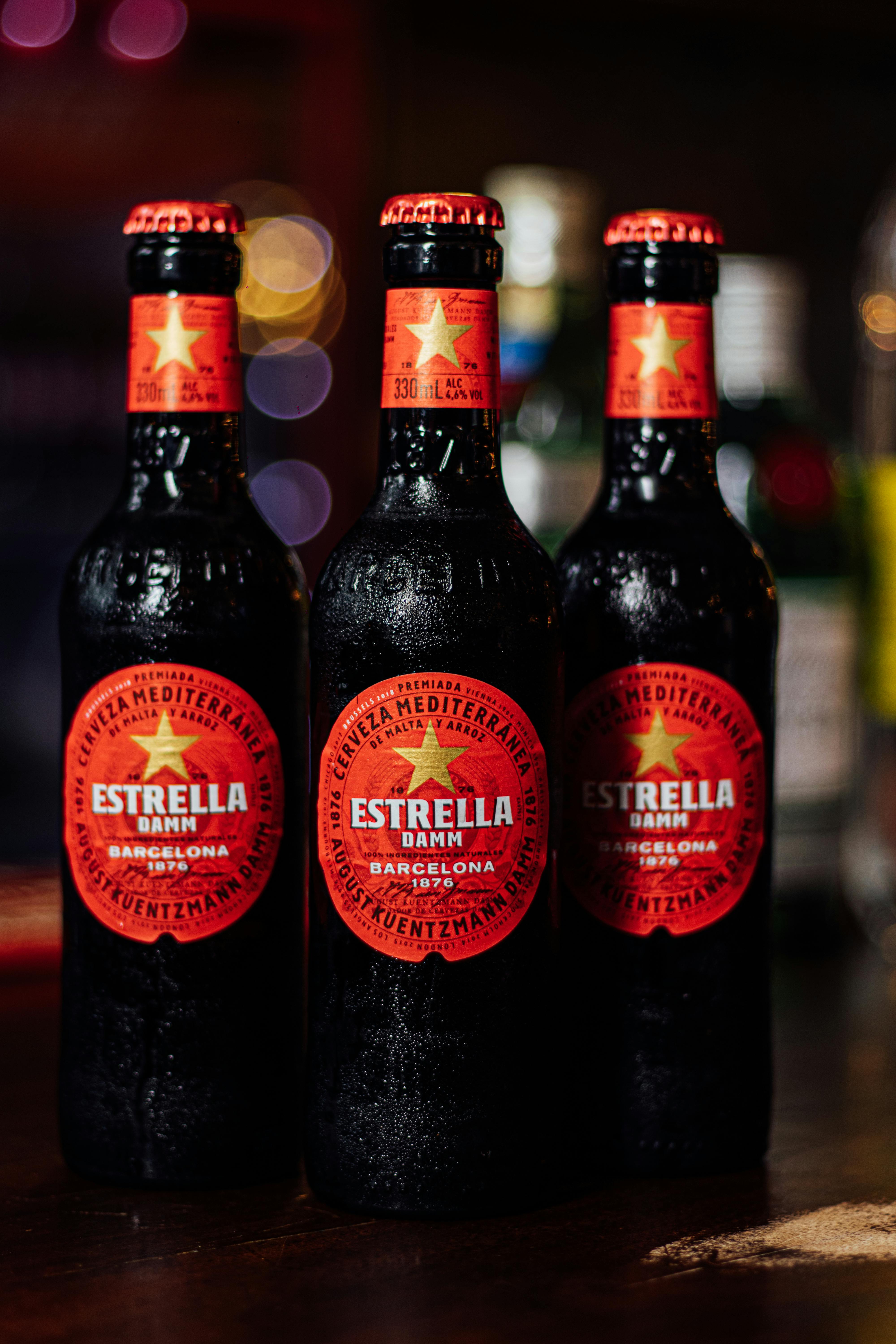 Close-up of three Estrella Damm beer bottles on a bar counter with blurred background.