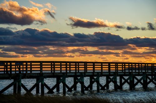 Free Silhouette of a wooden pier over the ocean against a vibrant sunset sky with clouds. Stock Photo