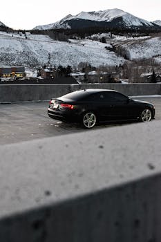 A sleek black car parked with a snowy mountain backdrop under a cloudy sky.