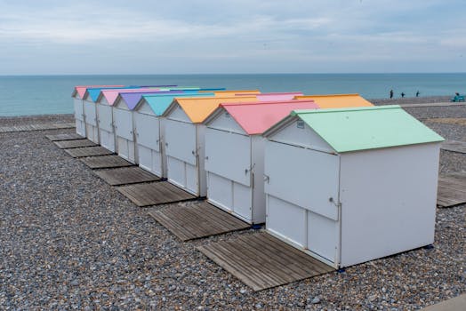 Vibrant beach huts with colorful roofs line a pebbled beach on a cloudy day.