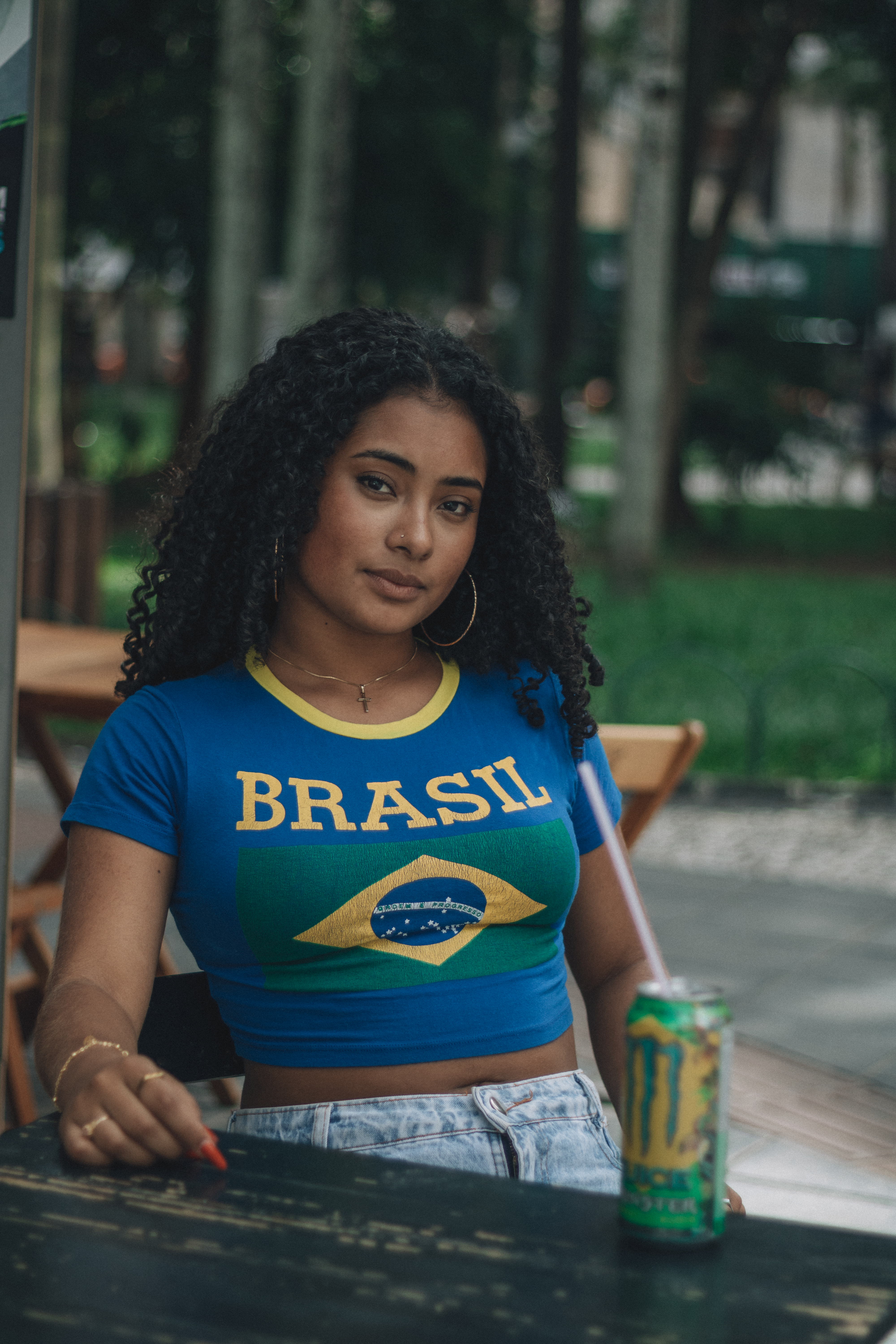 Young woman sitting outdoors wearing a Brazil T-shirt, holding a beverage can.