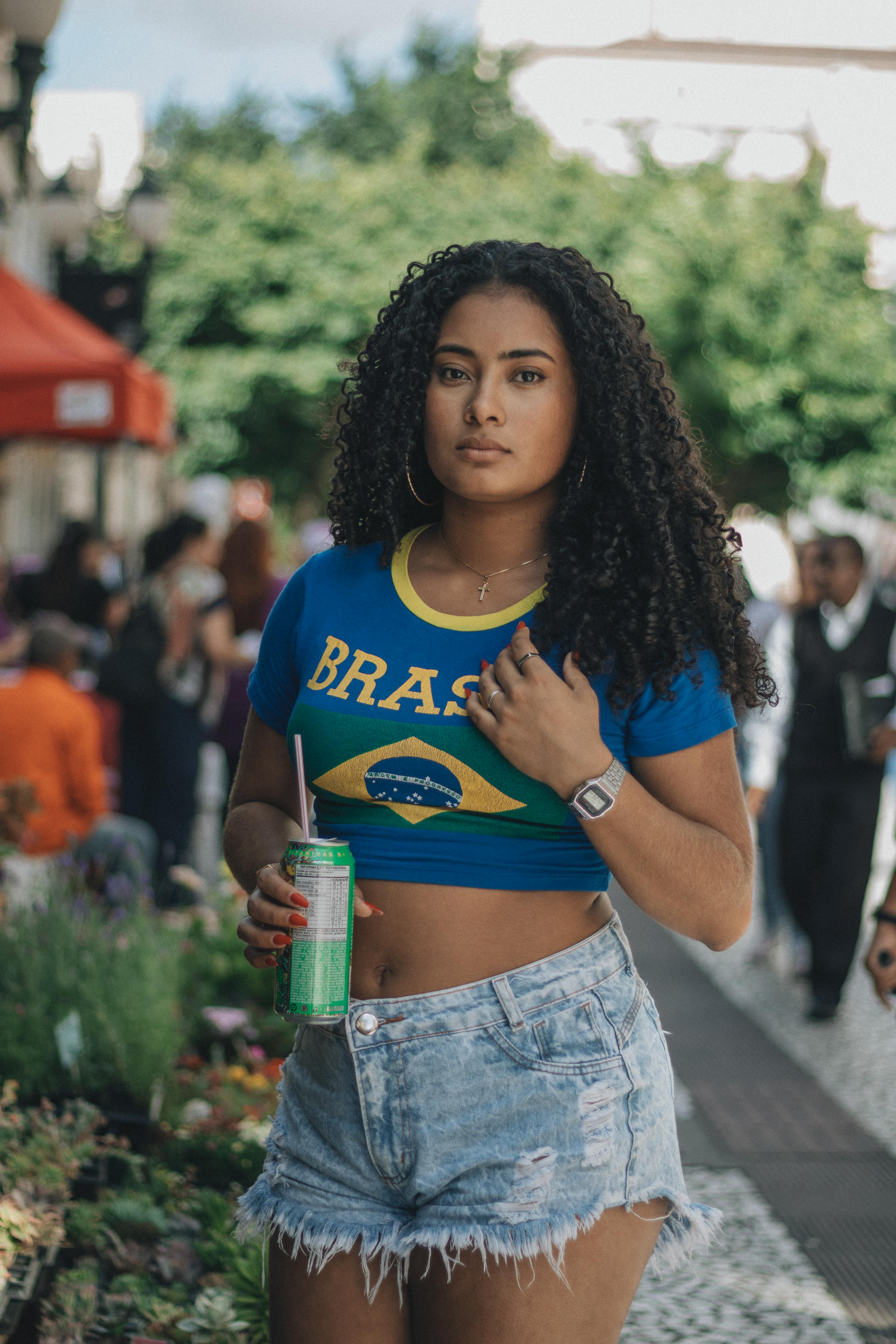 Woman in Brazilian flag shirt walking with a drink, showcasing summer fashion and confidence.