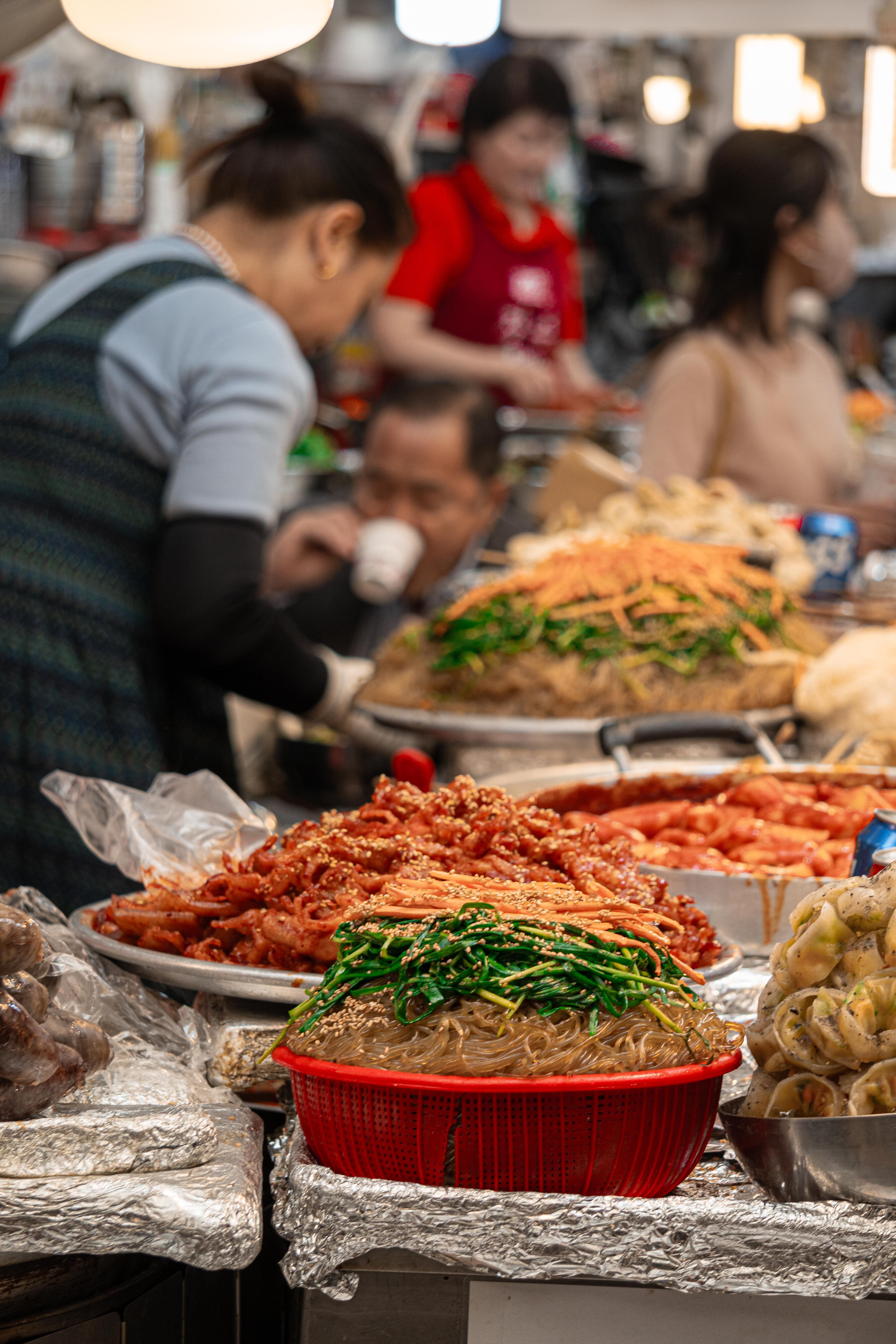 Colorful dishes at a bustling Asian market food stall with people in the background.