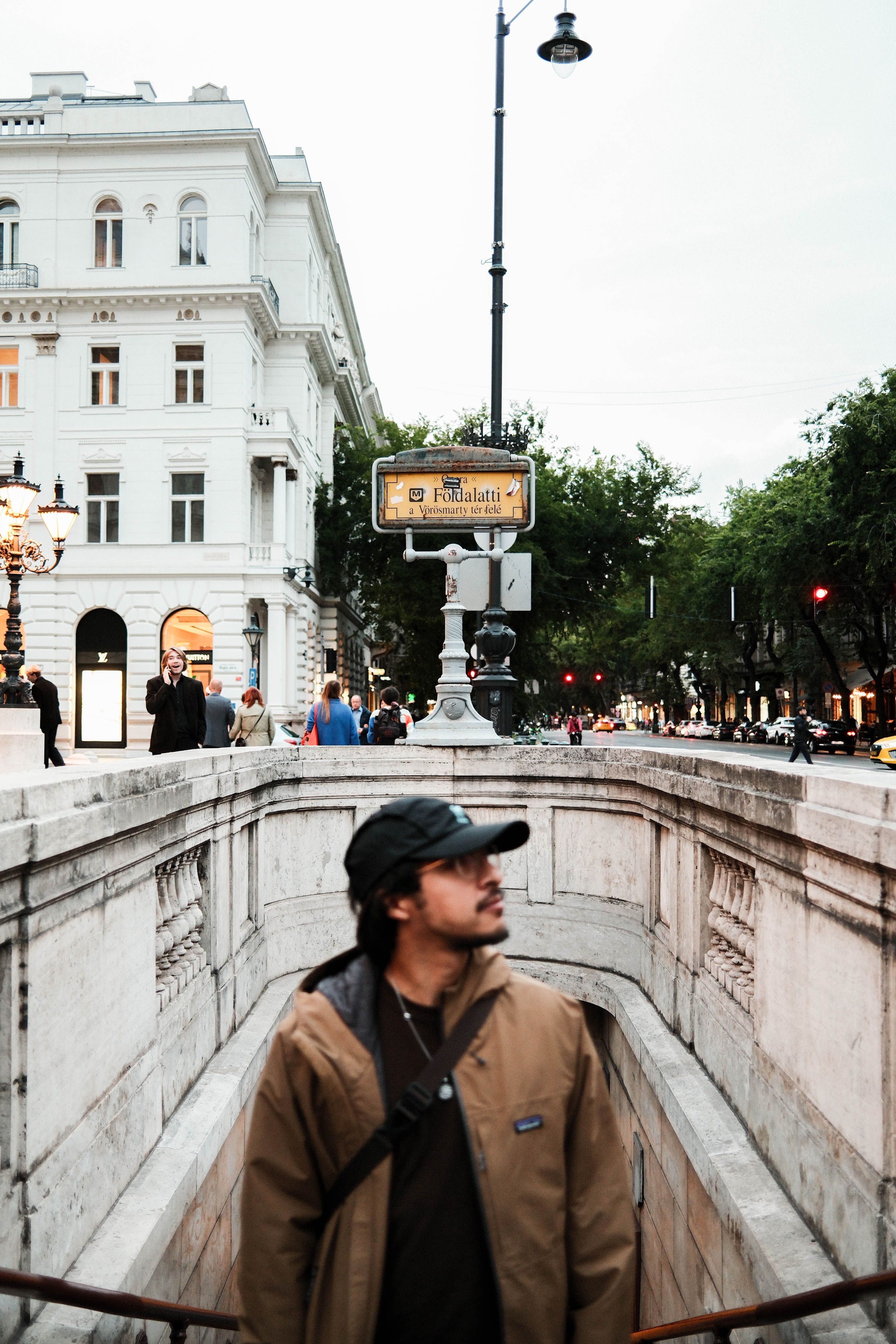 A man stands outside a metro entrance in Budapest, surrounded by historic architecture and city life.