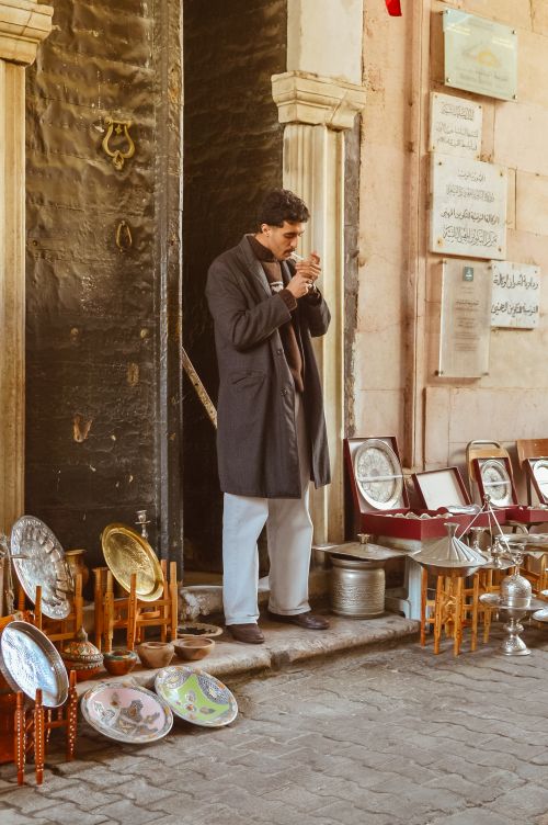 Free Man in coat standing outside a shop with metalware and plates on display. A scene from a traditional market. Stock Photo