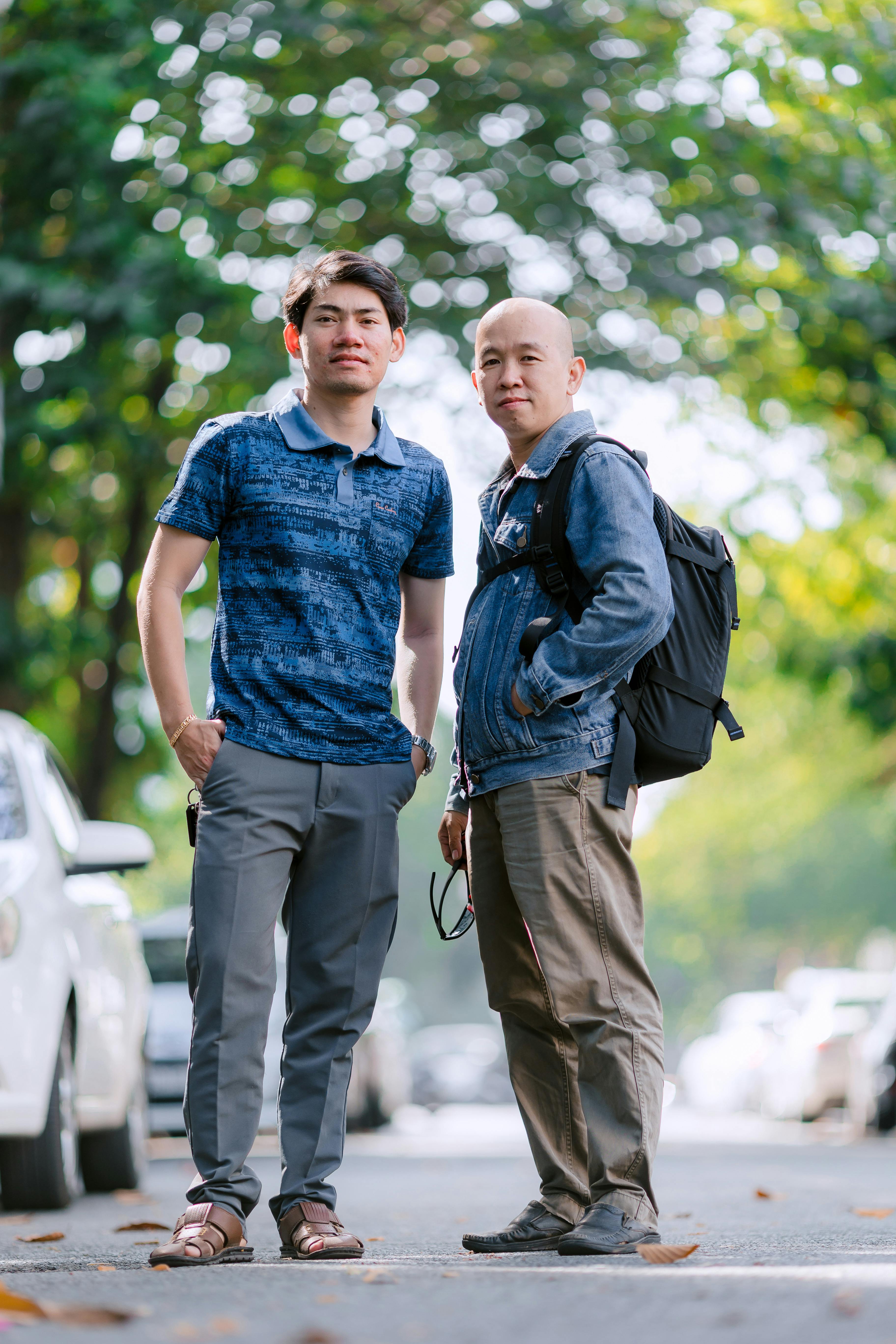 Two men standing confidently on a tree-lined street, with cars in the background.