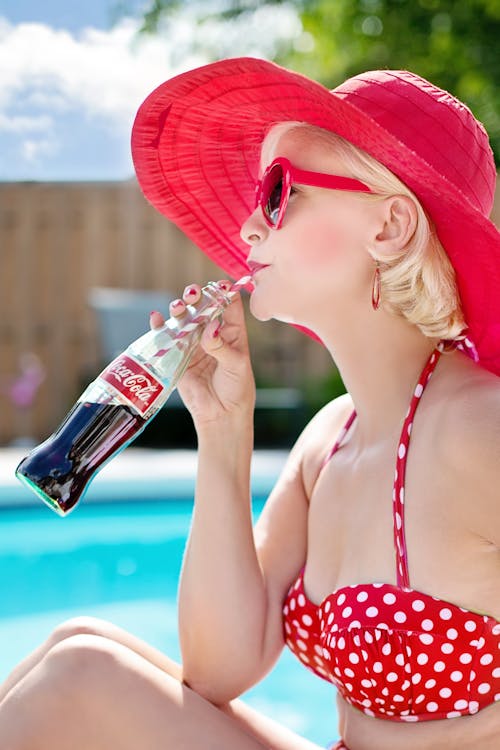 Vrouw In De Rode Bikini Van De Stipdruk Die Rode Kaderzonnebril Dragen Die De Drank Van Coca Cola In Blauwe En Witte Wolken Drinken