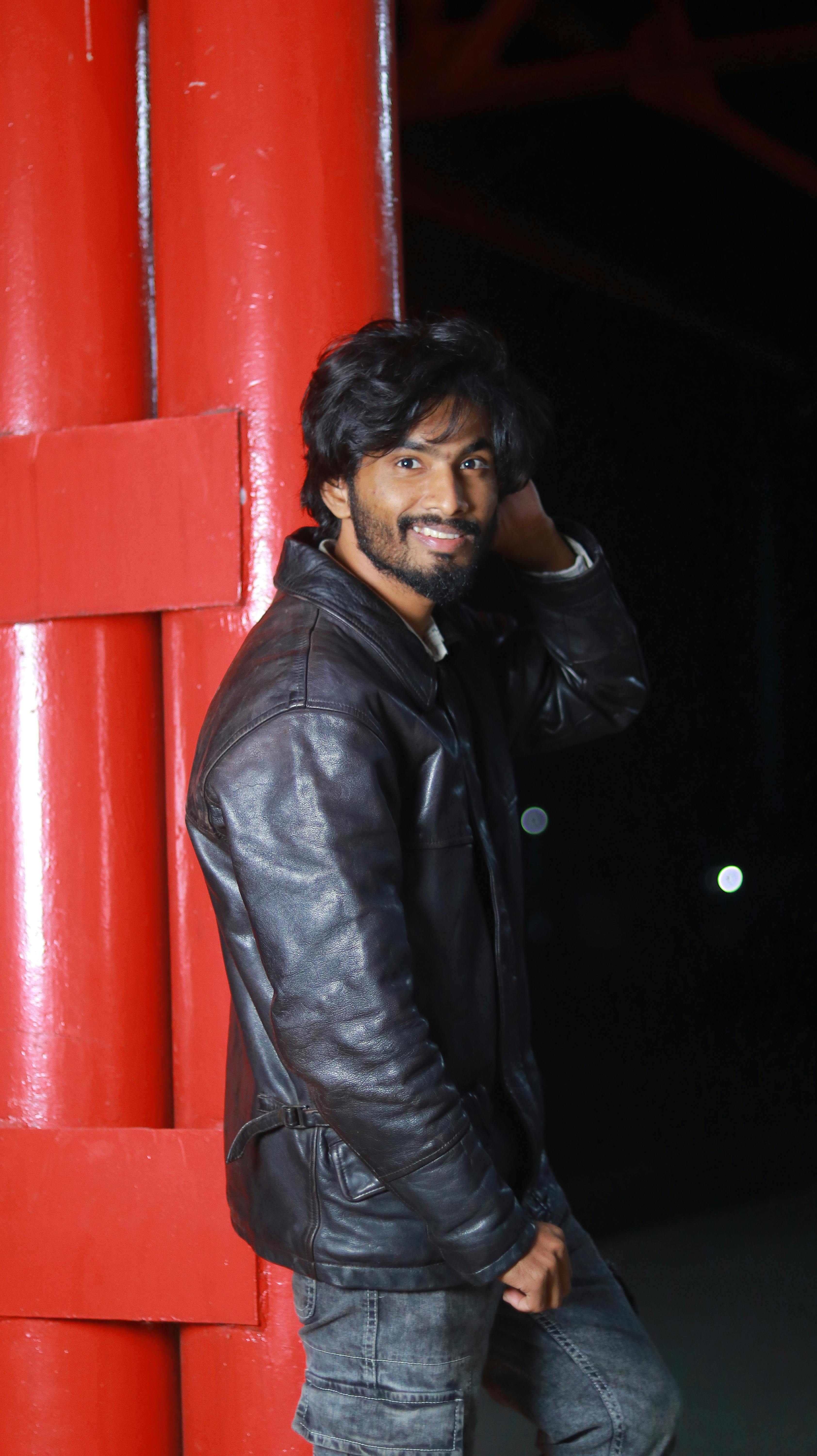 A young man poses casually against a red structure while wearing a leather jacket at night.