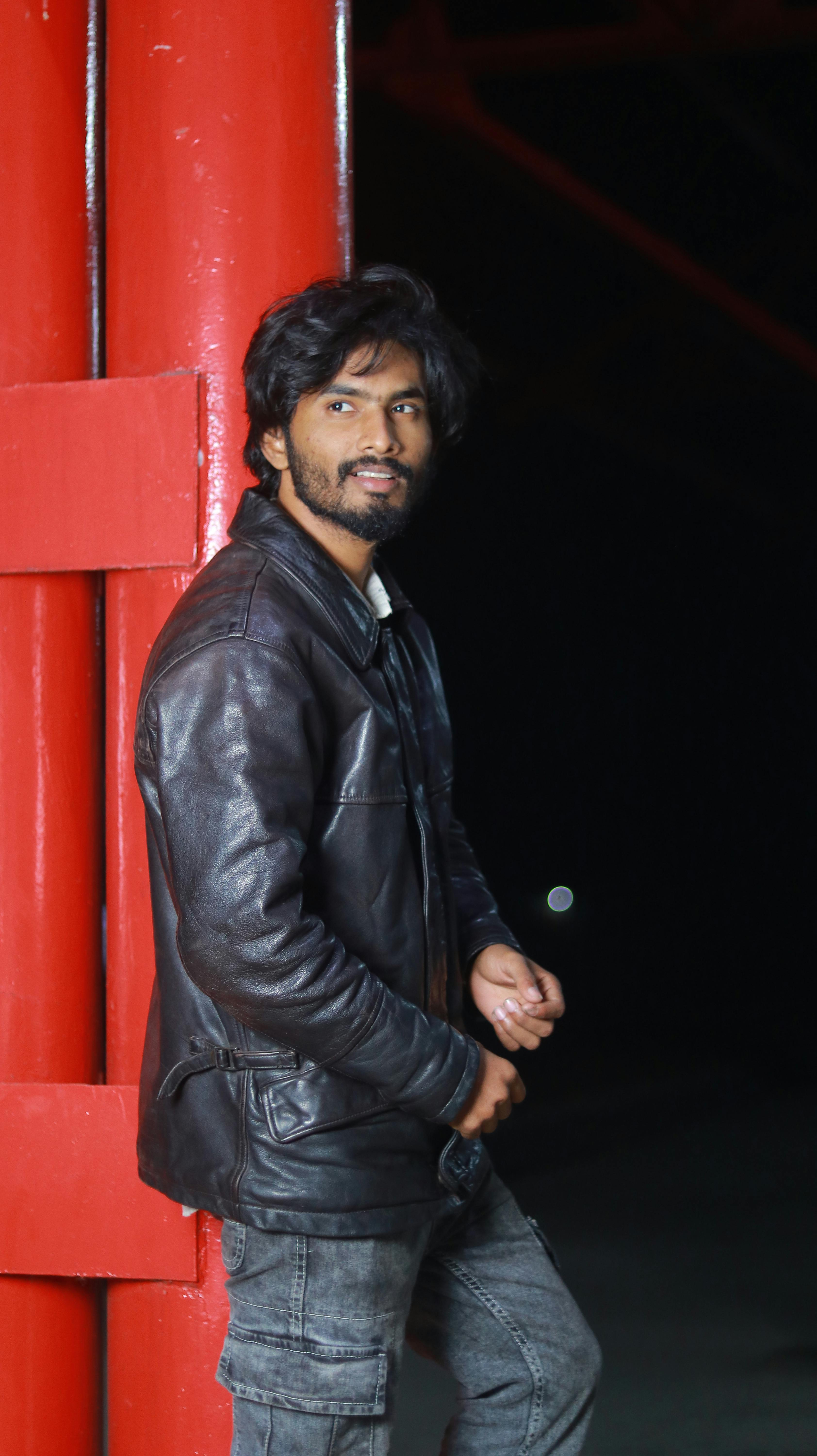 grátis Jovem de jaqueta de couro encostado em um pilar vermelho à noite. Foto profissional