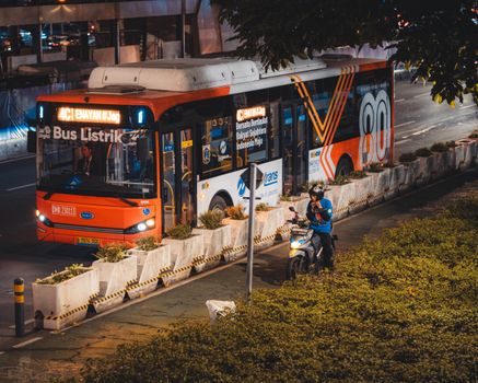 Electric bus and motorcyclist in vibrant Jakarta city at night.