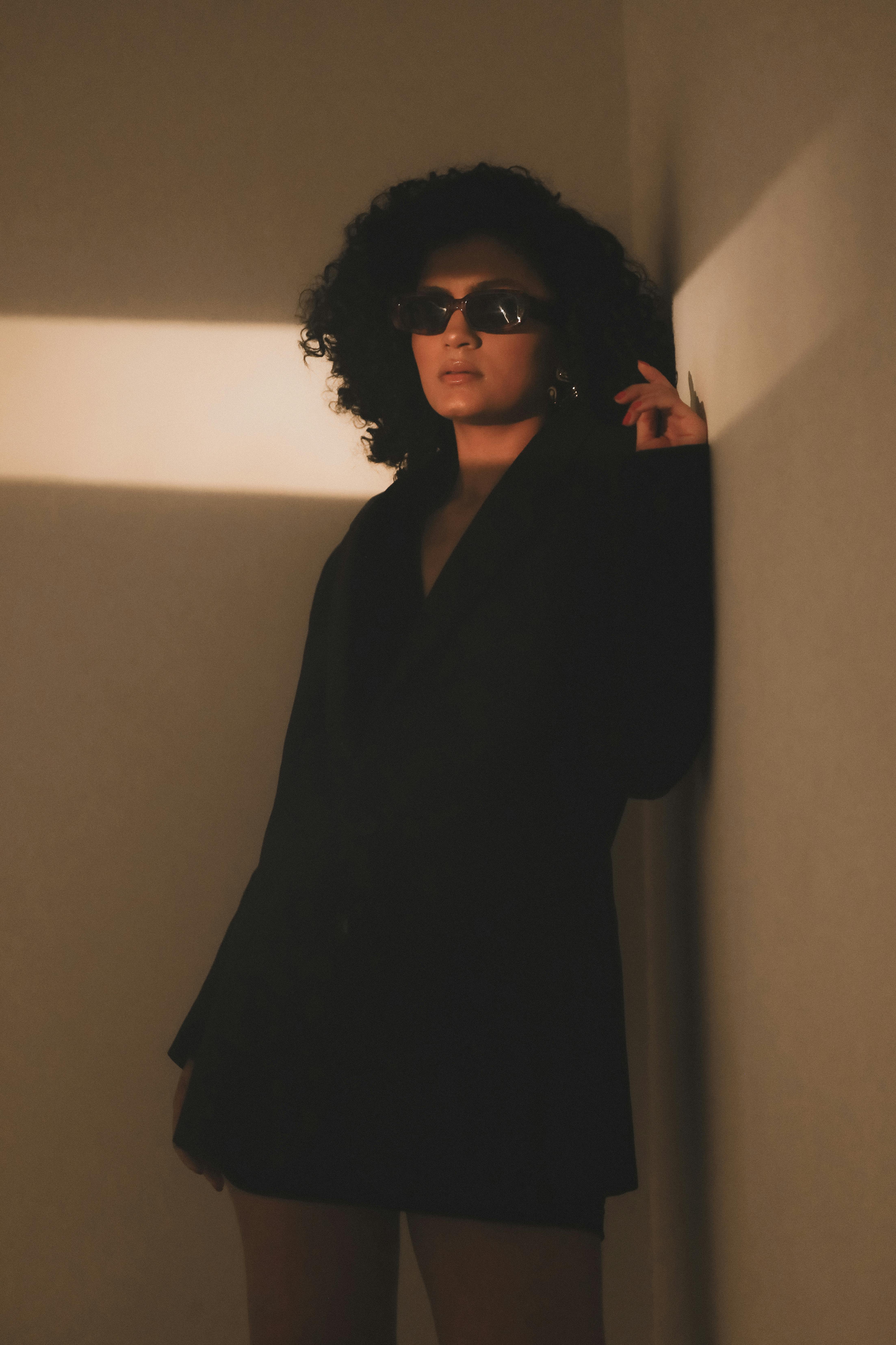 Chic woman wearing sunglasses and black outfit, backlit by warm light against a wall.