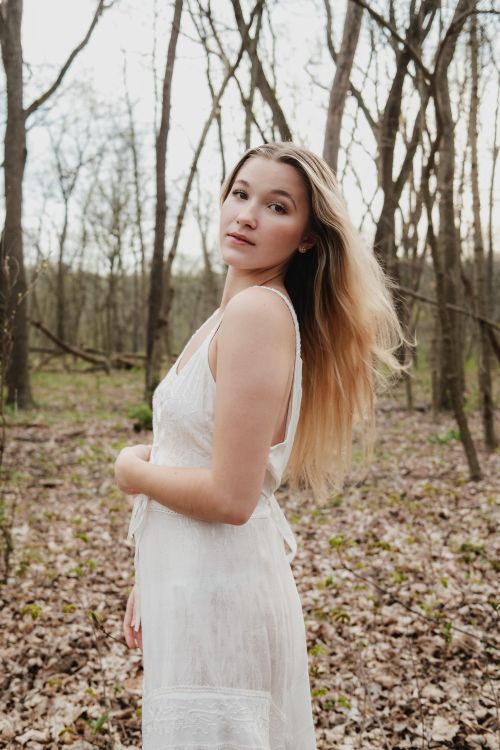 Free A young woman in a white dress stands elegantly in a serene forest setting. Stock Photo